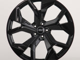 A black, glossy alloy wheel with a five-spoke design, displaying the Audi logo in the center, is positioned against a plain light gray background.