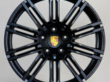 A black alloy wheel with multiple spokes features a central emblem displaying the Porsche logo. The wheel is positioned against a plain light gray background.
