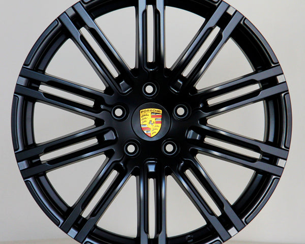 A black alloy wheel with multiple spokes features a central emblem displaying the Porsche logo. The wheel is positioned against a plain light gray background.