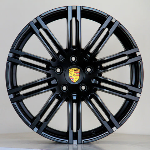 A black alloy wheel with multiple spokes features a central emblem displaying the Porsche logo. The wheel is positioned against a plain light gray background.