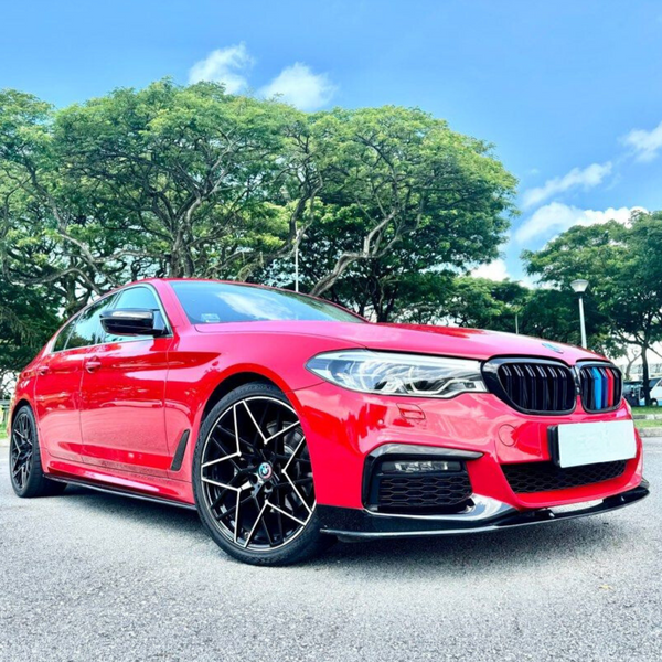 A red sedan with sleek black rims parked on a gray road, framed by lush green trees and a clear blue sky in the background.