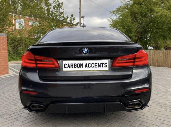 A black BMW sedan is parked on a paved driveway with trees and a brick building in the background. The license plate reads "CARBON ACCENTS."
