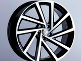A sleek, modern Volkswagen wheel rim featuring bold geometric spokes is displayed on a black pedestal against a neutral background. It has a glossy black and silver finish with the Volkswagen logo at the center.