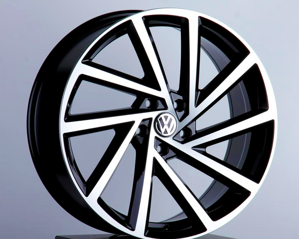 A sleek, modern Volkswagen wheel rim featuring bold geometric spokes is displayed on a black pedestal against a neutral background. It has a glossy black and silver finish with the Volkswagen logo at the center.