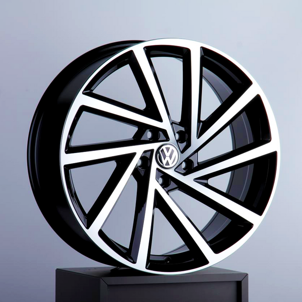 A sleek, modern Volkswagen wheel rim featuring bold geometric spokes is displayed on a black pedestal against a neutral background. It has a glossy black and silver finish with the Volkswagen logo at the center.