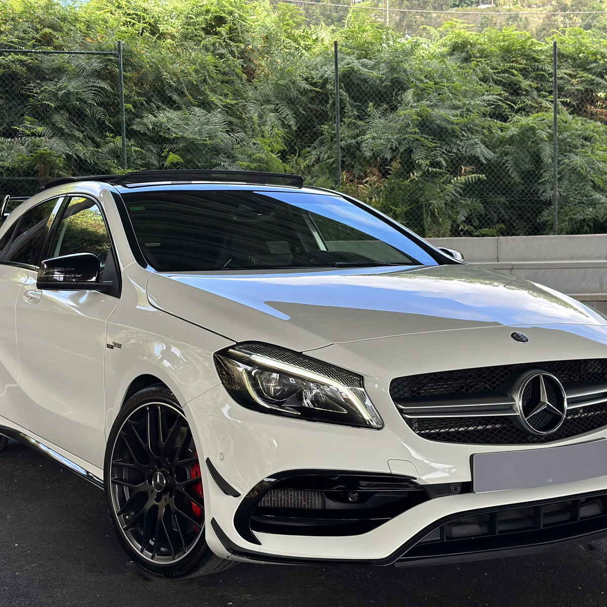 A white Mercedes-Benz car, stationary, is parked on black asphalt in front of a chain-link fence with dense greenery behind it.