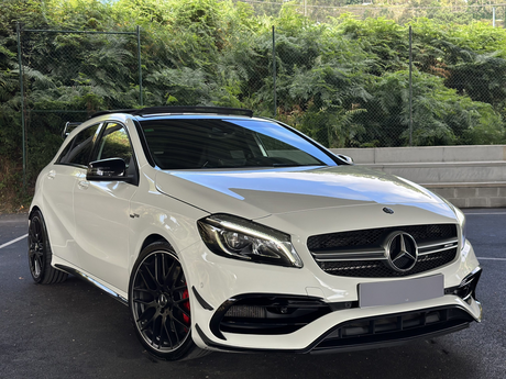 A white Mercedes-Benz car, stationary, is parked on black asphalt in front of a chain-link fence with dense greenery behind it.