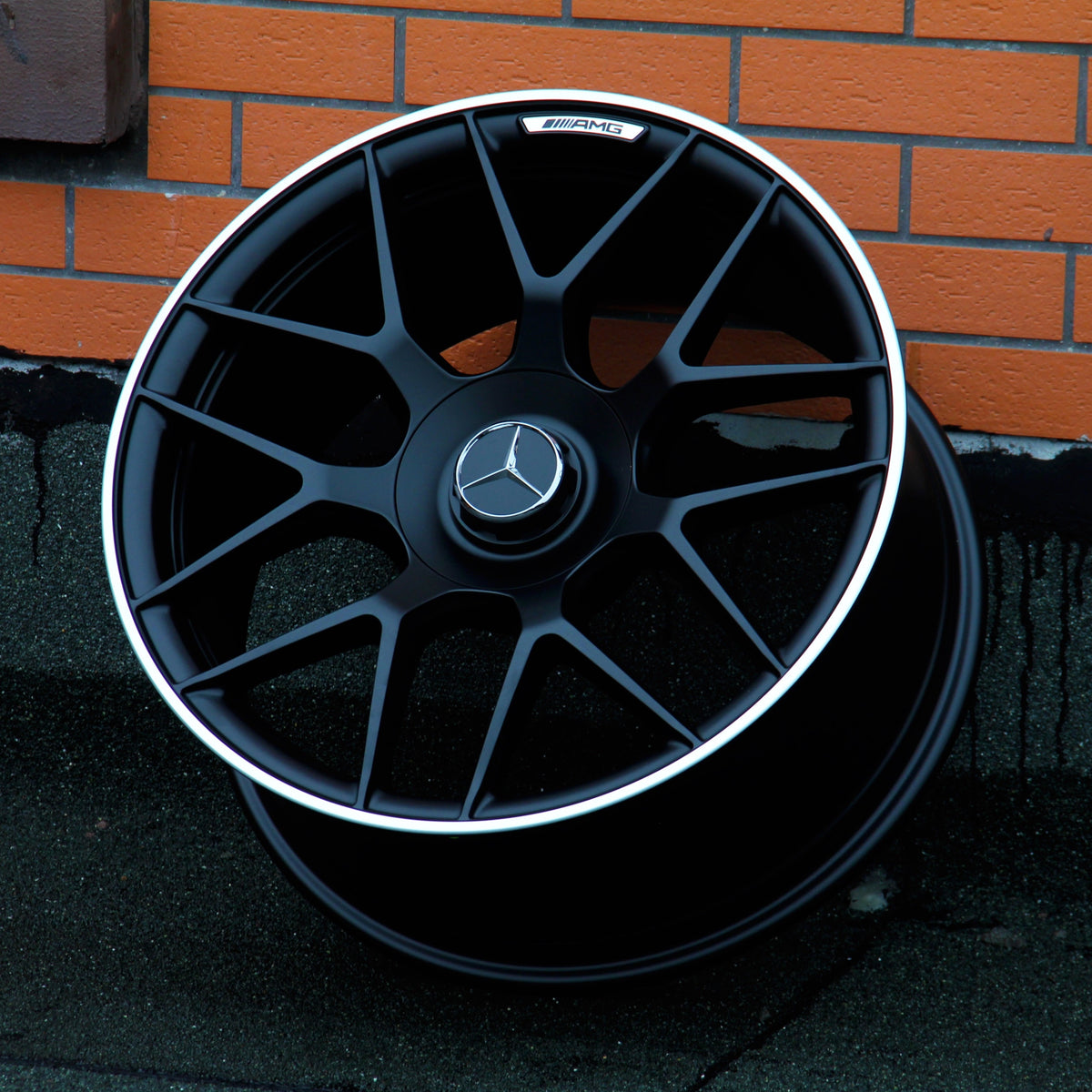 A black Mercedes-Benz AMG wheel leans against an orange brick wall. The wheel features a silver rim and the Mercedes emblem at its center.