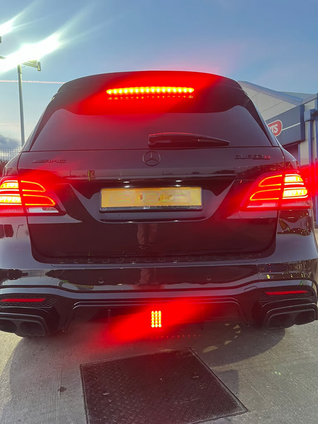 A black Mercedes parked with brake lights illuminated; the setting is an outdoor area near a building labeled "ATS." The brand and "AMG" are visible on the rear.