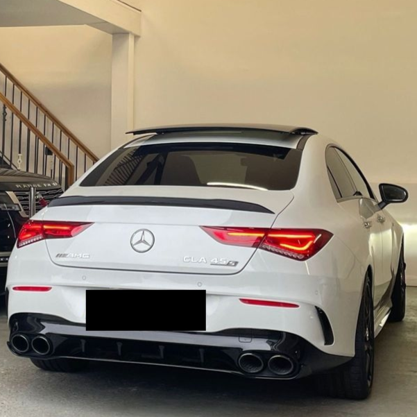 A white Mercedes-Benz AMG CLA 45 S is parked in a garage, illuminated by indoor lighting. It features quad exhausts, tinted windows, and visible branding. Text: "AMG" and "CLA 45 S".