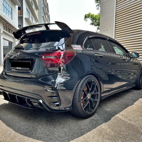 A black Mercedes hatchback, parked beside industrial buildings, features dark-tinted windows, a rear spoiler, and alloys. Reflective surfaces display building textures. Visible logos: "AMG" and "Ecobenzo."