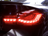 A car's taillight is illuminated in bright red, showing a sleek, modern design with horizontal light strips, set against a glossy black car exterior.