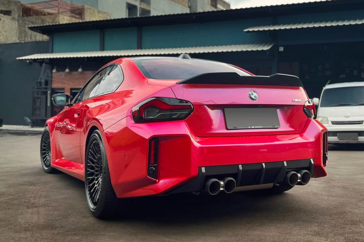 A red BMW M2 car is parked on a concrete surface, facing away, showing its sleek rear design and quad exhausts. It's set against an industrial building backdrop.