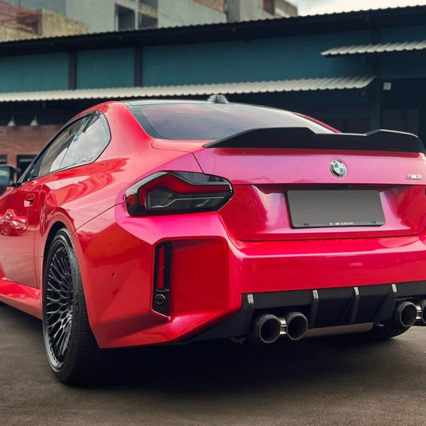 A red BMW M2 car is parked on a concrete surface, facing away, showing its sleek rear design and quad exhausts. It's set against an industrial building backdrop.