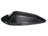 A carbon fiber cover with a sleek, aerodynamic shape sits isolated against a plain white background, highlighting its glossy weave pattern.