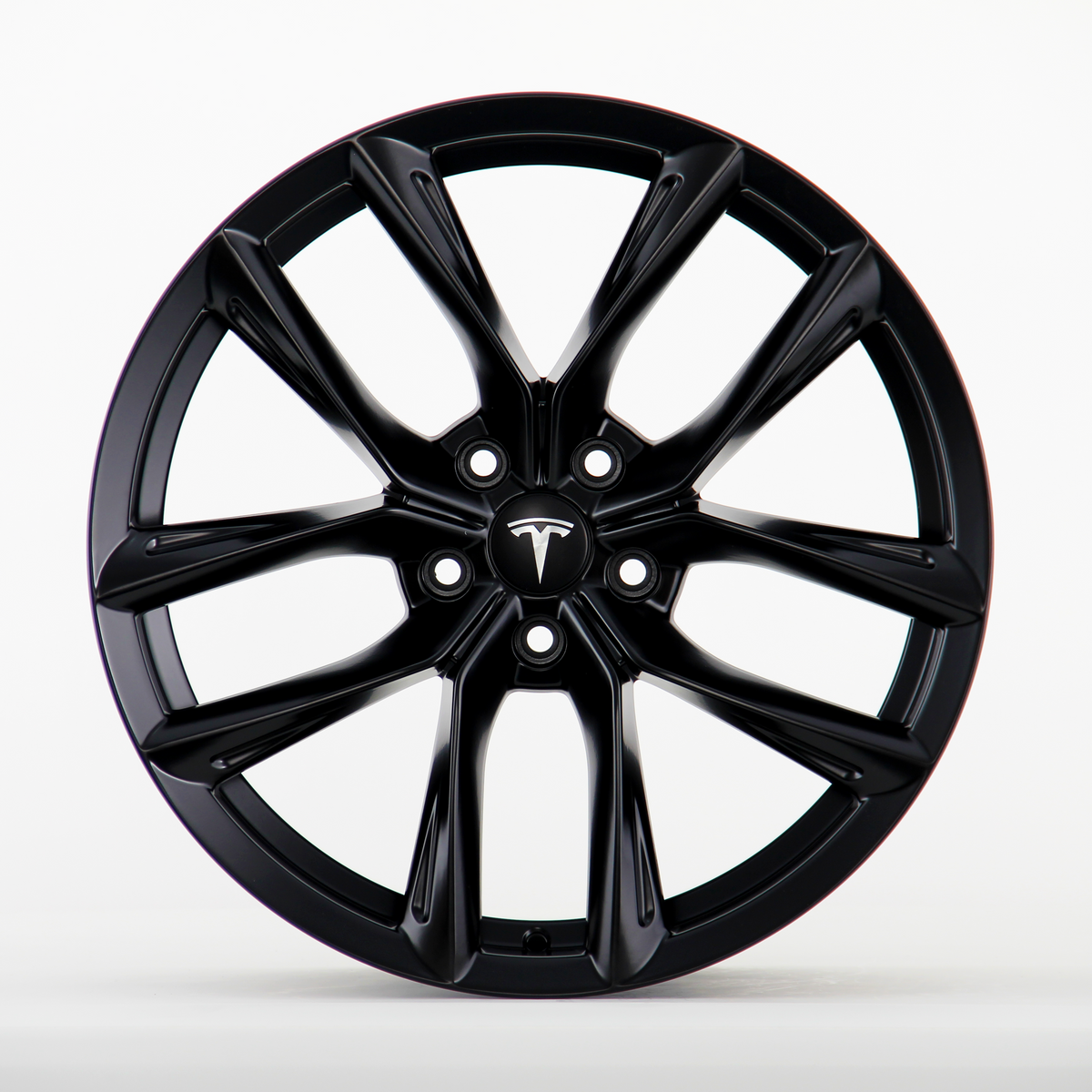 A black Tesla wheel rim with a sleek, multi-spoke design is shown in a clean, white studio setting. The central hub cap features the Tesla logo prominently.
