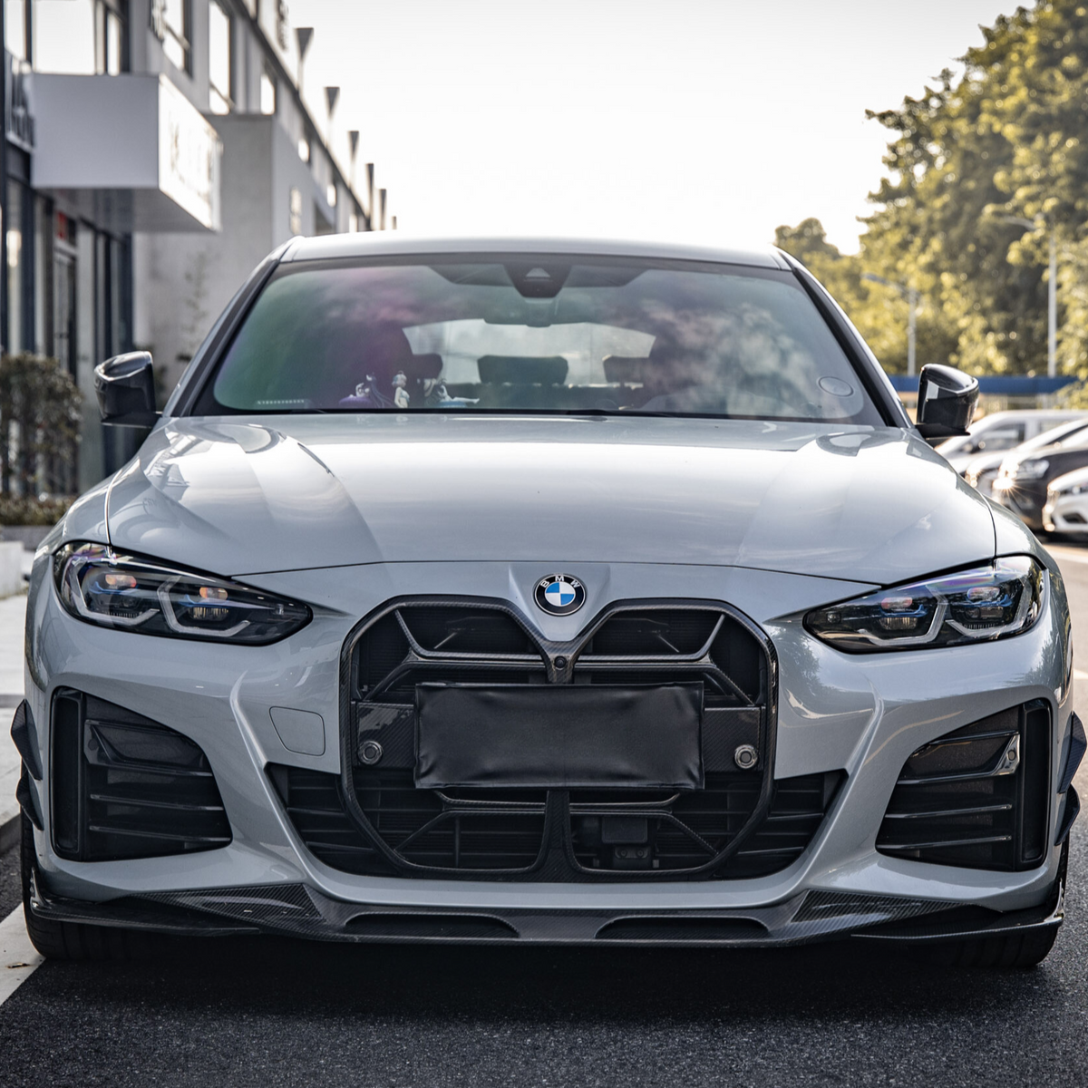A silver car with a prominent grille is parked on a street beside a line of buildings, with trees visible in the background under a clear sky.