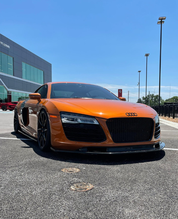 An orange sports car with sleek curves and black rims is parked in a lot near a modern building. The sky is clear, and other cars are in the distance.