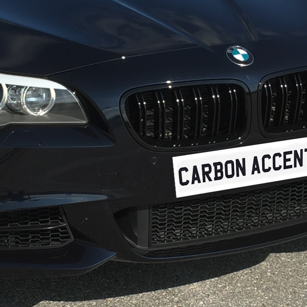 A black car features carbon accents on its front grille, parked on a paved surface. Text on the license plate area reads "CARBON ACCENTS."