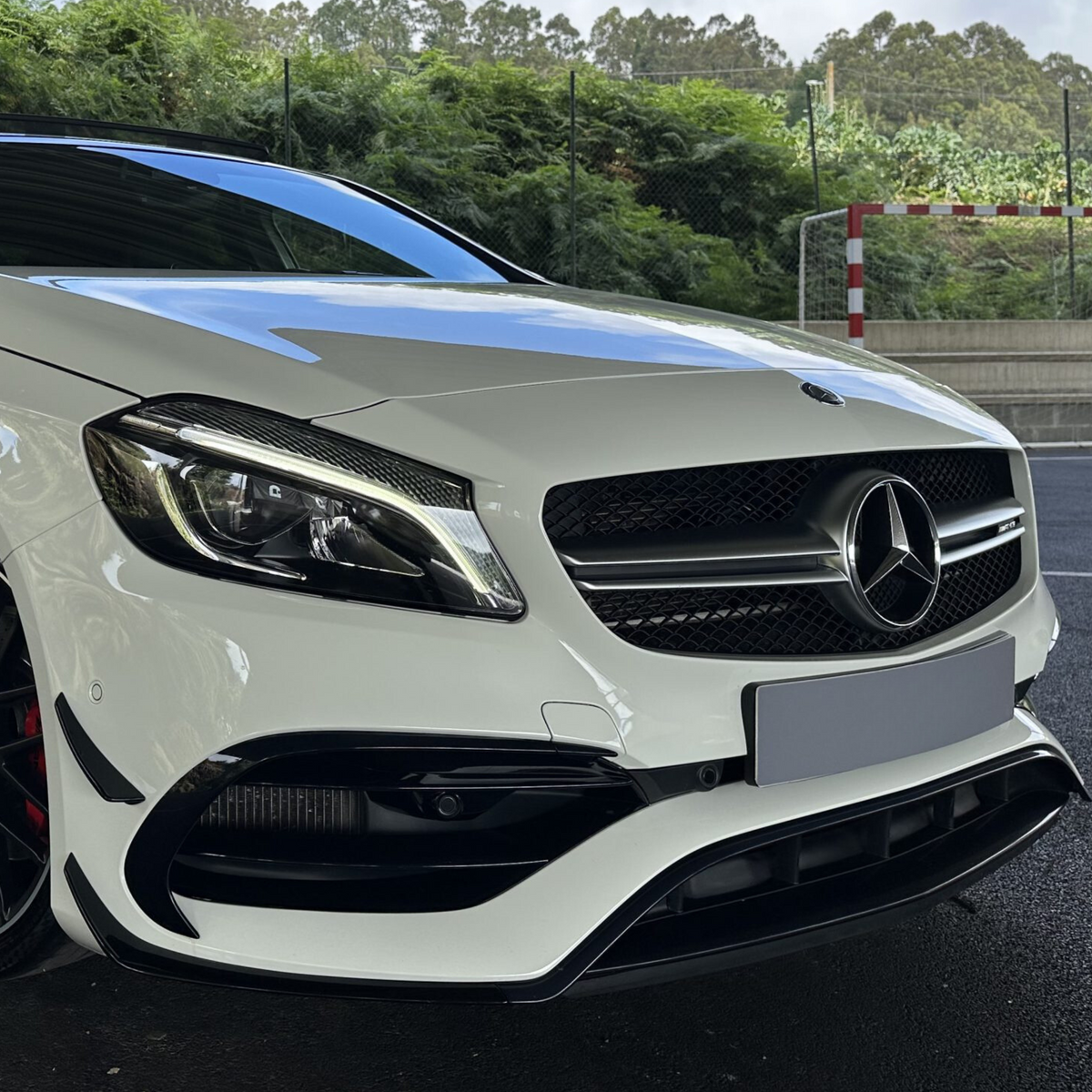 A white car with a prominent front grille and sleek headlights is parked outdoors, near a fence and green bushes in the background.