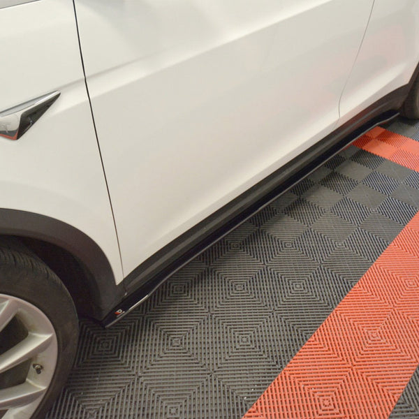 A white car is parked, showcasing sleek side skirts, on a checkered floor composed of red and gray tiles in a garage setting.