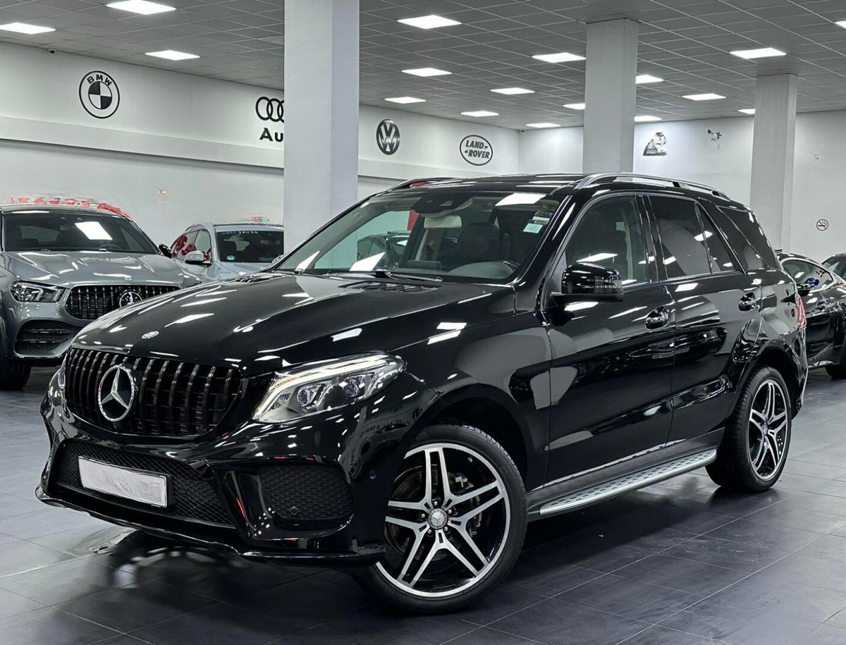 A black Mercedes SUV is parked inside a car showroom, surrounded by other vehicles. Logos of BMW, Audi, Volkswagen, and Land Rover are displayed on the walls.