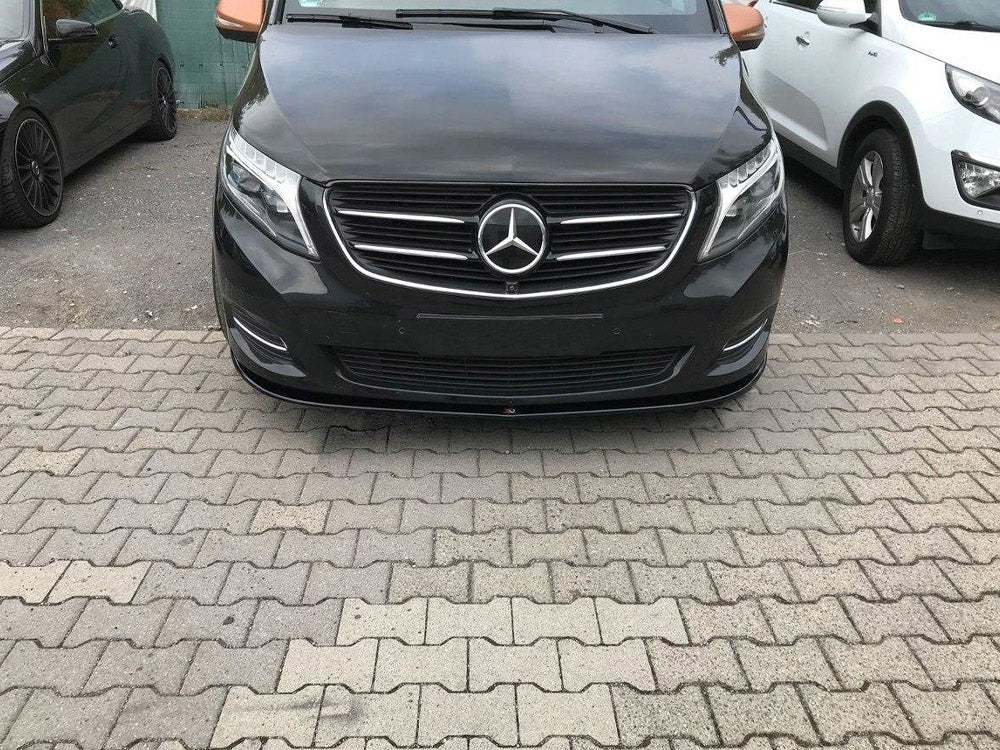 A black Mercedes-Benz van is parked on a paved area, surrounded by other cars, including a white SUV on the right and a dark vehicle on the left.
