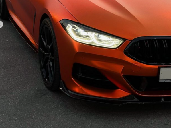 Car's front end with bright headlights illuminated; matte orange finish and black grille. Parked on a dark asphalt surface, displaying a sporty and sleek design.