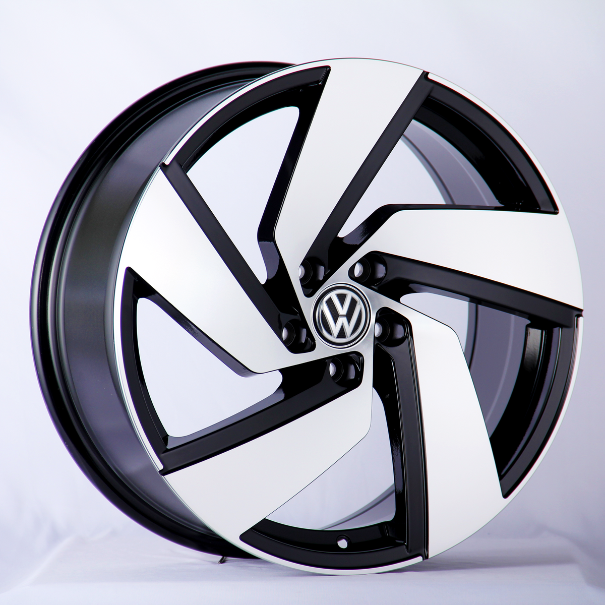 A car wheel rim displays a black and silver geometric design featuring a central Volkswagen logo. It is positioned upright against a neutral, softly lit background.