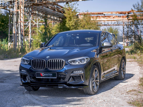 A black BMW SUV is parked on a gravel path, with metal scaffolding and green foliage in the background. The license plate reads "MAXTON design."