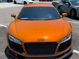 Orange Audi sports car parked in a lot, surrounded by several other high-end cars, including a white and a gray vehicle, outside a glass-fronted building.