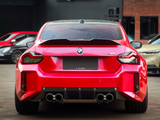 A bright red BMW M2 car is parked, displaying quad exhaust pipes. It is surrounded by other parked vehicles in an outdoor setting. Text on plate reads, 