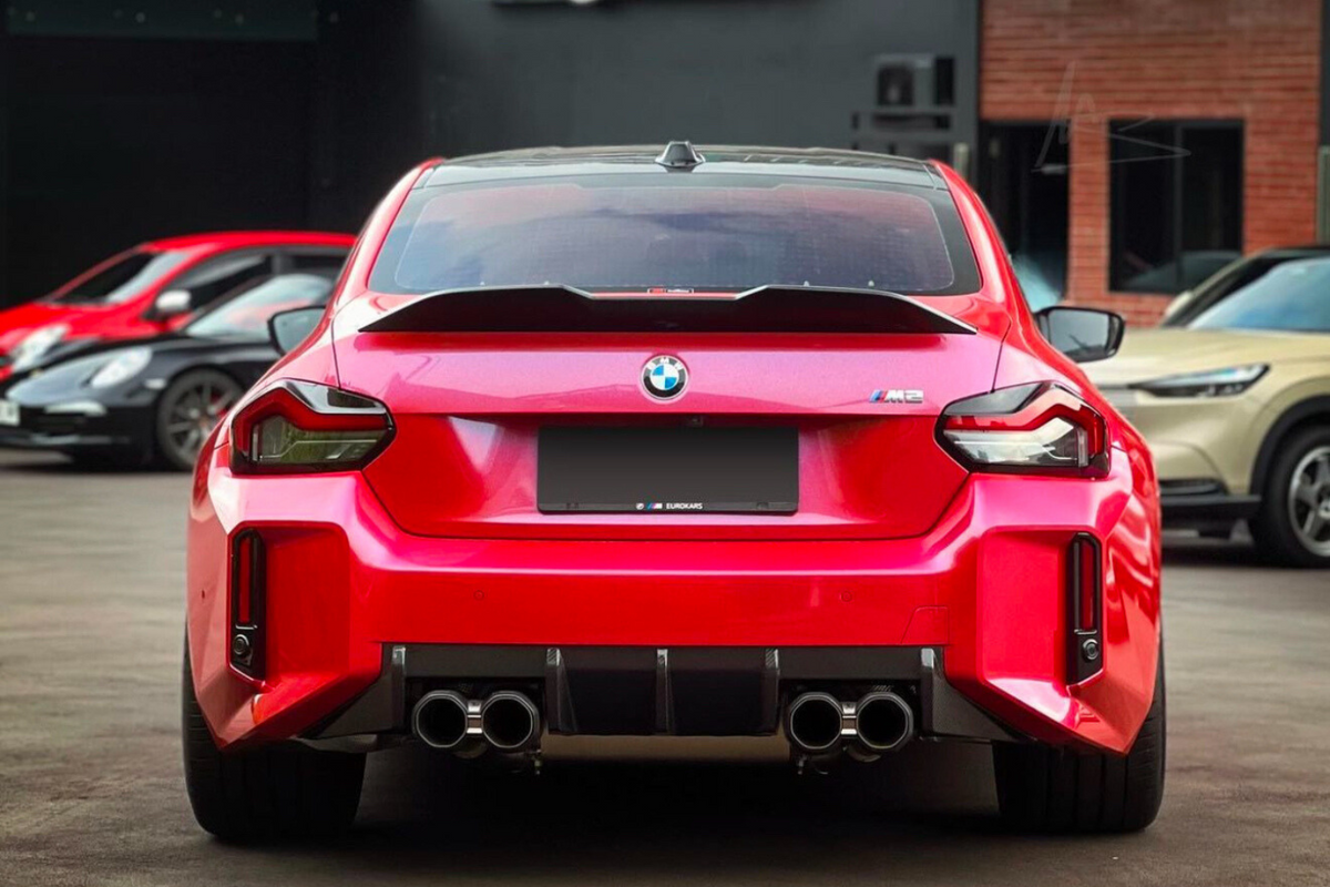 A bright red BMW M2 car is parked, displaying quad exhaust pipes. It is surrounded by other parked vehicles in an outdoor setting. Text on plate reads, "EUROKARS."