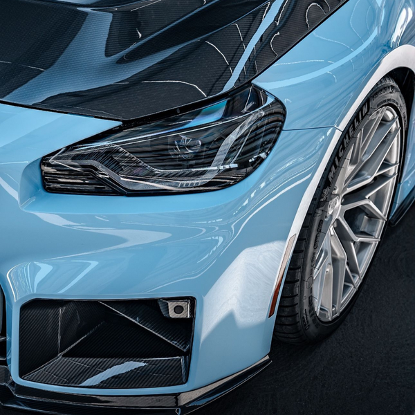 A light blue car's sleek front view, showcasing a detailed headlight, carbon fiber elements, and a Michelin tire in a glossy, clean indoor setting.