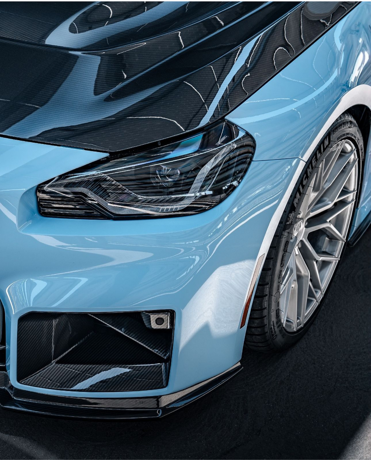 A light blue car's sleek front view, showcasing a detailed headlight, carbon fiber elements, and a Michelin tire in a glossy, clean indoor setting.