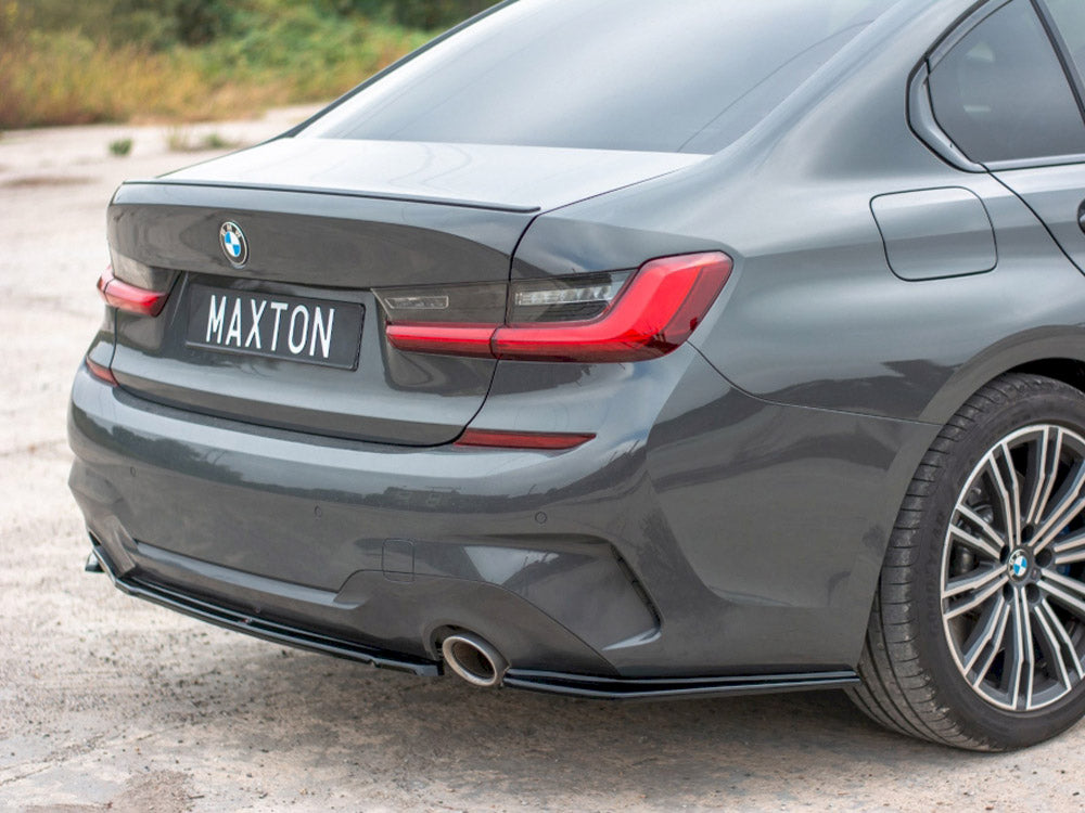 A grey BMW car is parked, showcasing its rear with a "MAXTON" license plate. The setting is an open, paved outdoor area with greenery in the background.