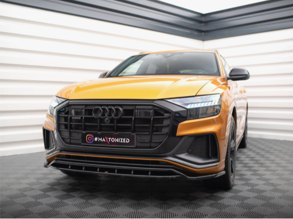 A gold-colored Audi SUV is parked on a checkered floor inside a showroom. The front grille features a prominent Audi logo. The license plate reads: "@MAXTONIZED."