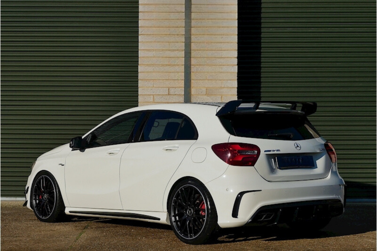 A white Mercedes-Benz AMG hatchback, parked, sits on gravel in front of large closed green garage doors. The car features a prominent black spoiler and alloy wheels.