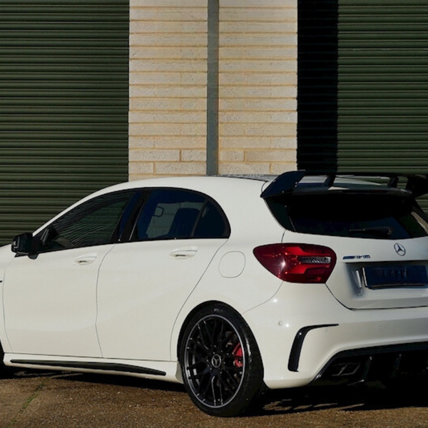 A white Mercedes-Benz AMG hatchback, parked, sits on gravel in front of large closed green garage doors. The car features a prominent black spoiler and alloy wheels.