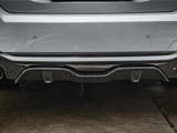 A car's rear bumper with dual exhaust pipes and a carbon fiber diffuser is parked on wet pavement, surrounded by a gray urban environment.