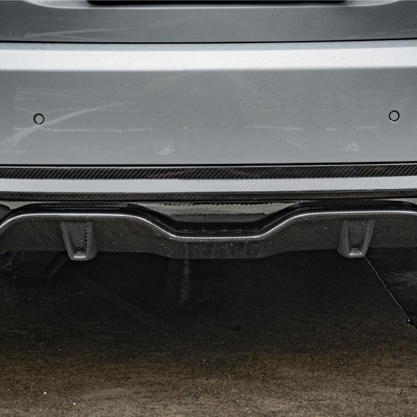 A car's rear bumper with dual exhaust pipes and a carbon fiber diffuser is parked on wet pavement, surrounded by a gray urban environment.