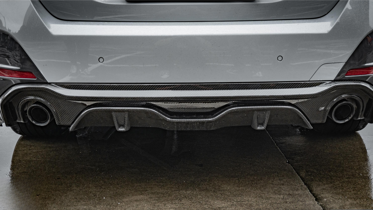 A car's rear bumper with dual exhaust pipes and a carbon fiber diffuser is parked on wet pavement, surrounded by a gray urban environment.