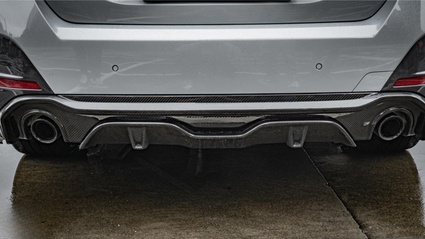 A car's rear bumper with dual exhaust pipes and a carbon fiber diffuser is parked on wet pavement, surrounded by a gray urban environment.