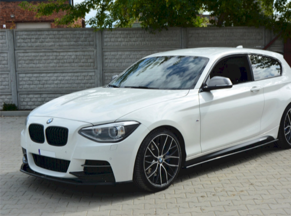 A white BMW car is parked on a paved surface. It features black rims and is positioned near a brick wall with greenery visible in the background.