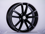 A black alloy wheel with a Volkswagen logo in the center, standing upright on a plain, light-colored background.
