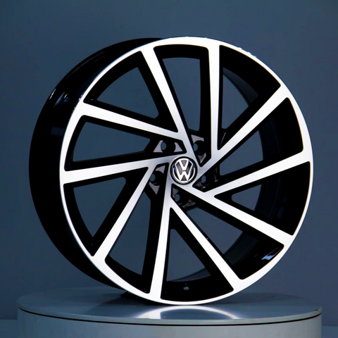 A black and silver car wheel with a geometric spoke design, displaying a Volkswagen logo in the center, is positioned upright on a reflective surface within a gray environment.