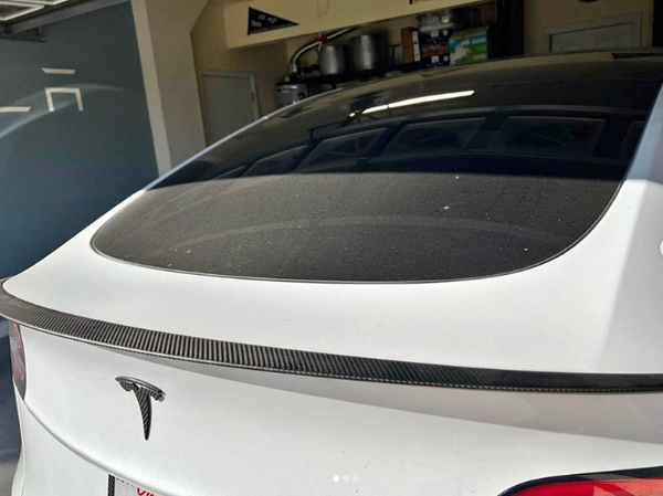 A white Tesla car is parked in a garage. The view shows the rear window and trunk, highlighted by a carbon fiber spoiler. Shelving with various items is visible in the background.
