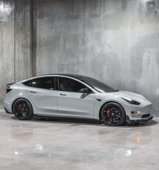 A sleek, gray sedan is parked on a shiny concrete floor, with black wheels and red brake calipers, set against an industrial, textured gray wall.