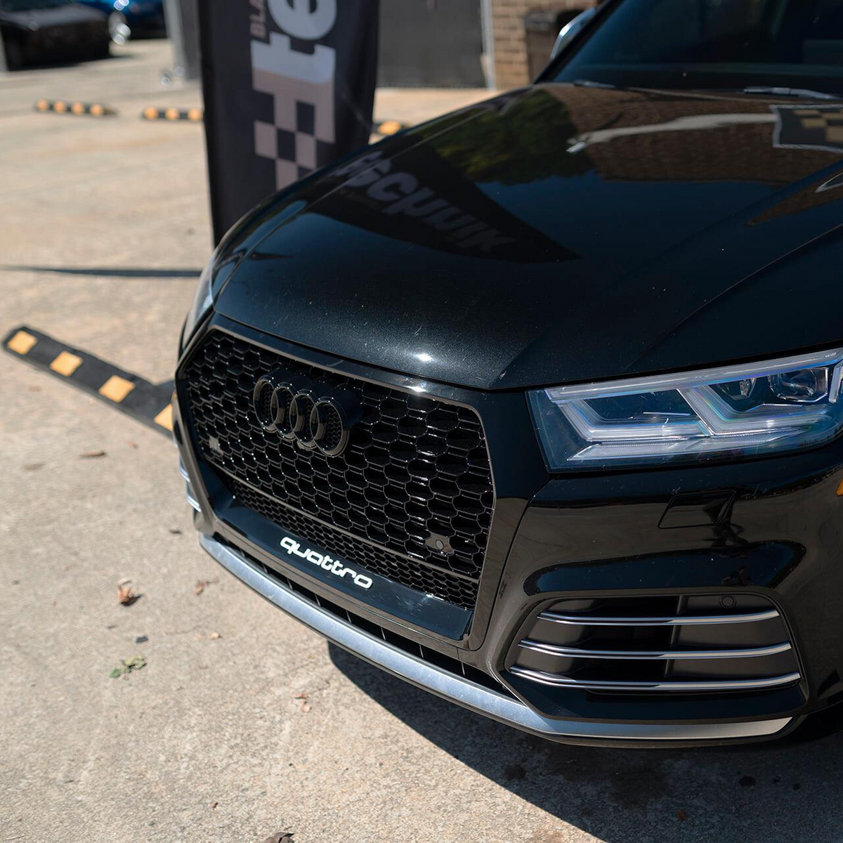 A black car displays a prominent grille with the Audi logo and "quattro" text, parked on a concrete surface near a vertical banner with partial text "te" visible.