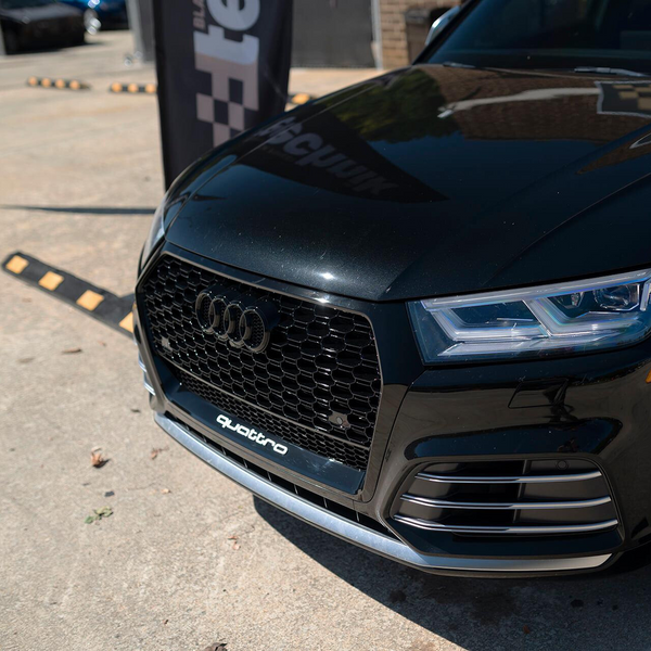 A black car displays a prominent grille with the Audi logo and "quattro" text, parked on a concrete surface near a vertical banner with partial text "te" visible.